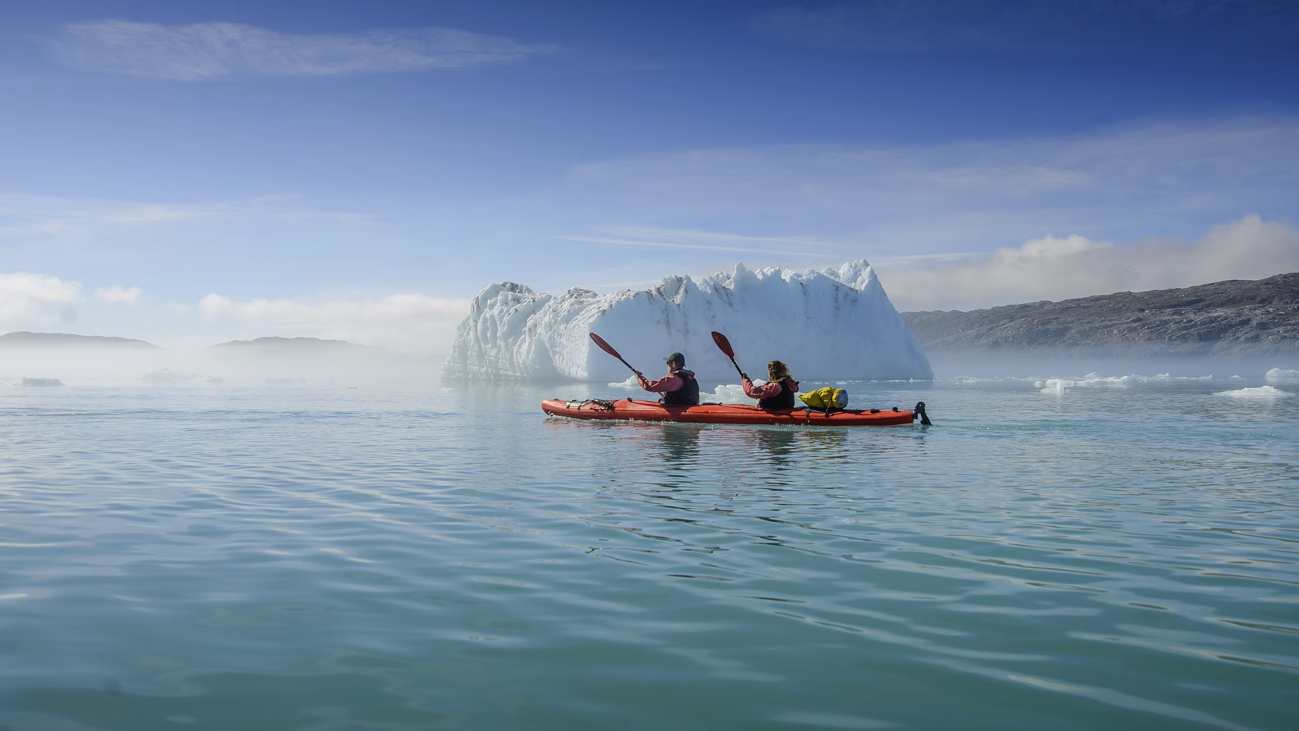 Two people kayaking on calm water near a large iceberg and rocky shore under a clear sky.