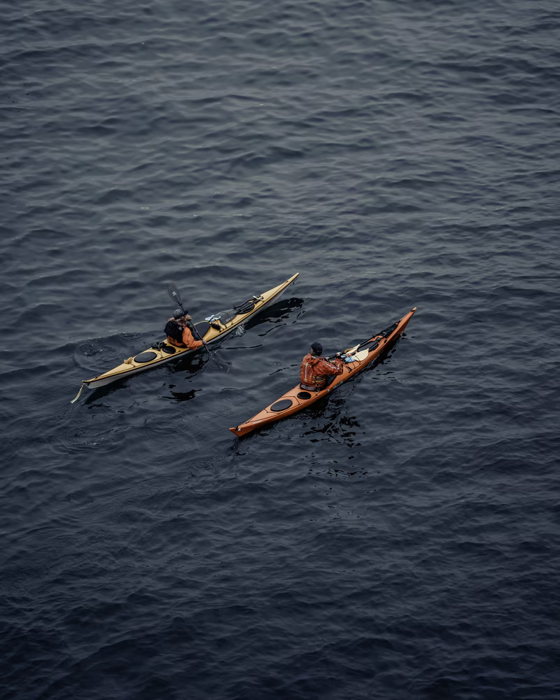 Two people kayaking on dark, rippled water wearing orange jackets and black helmets.