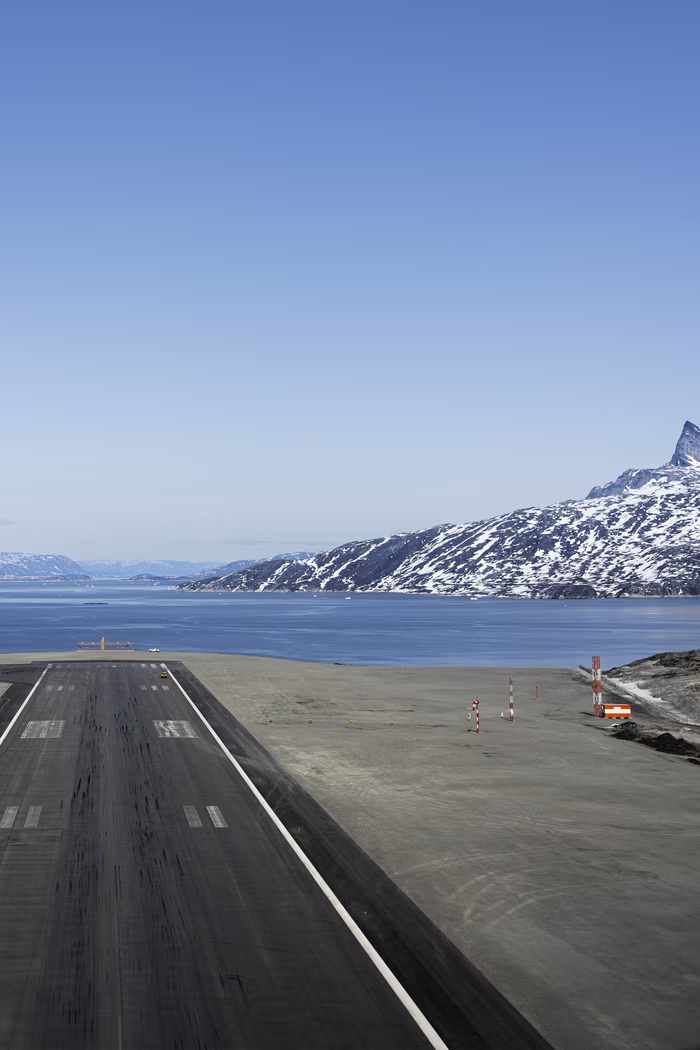 Aerial view of a runway beside a body of water with snow-capped mountains in the background under a clear sky.