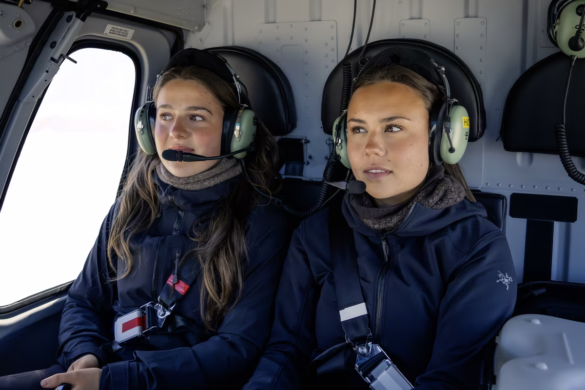 Two women wearing headsets and jackets sitting inside a helicopter cockpit.