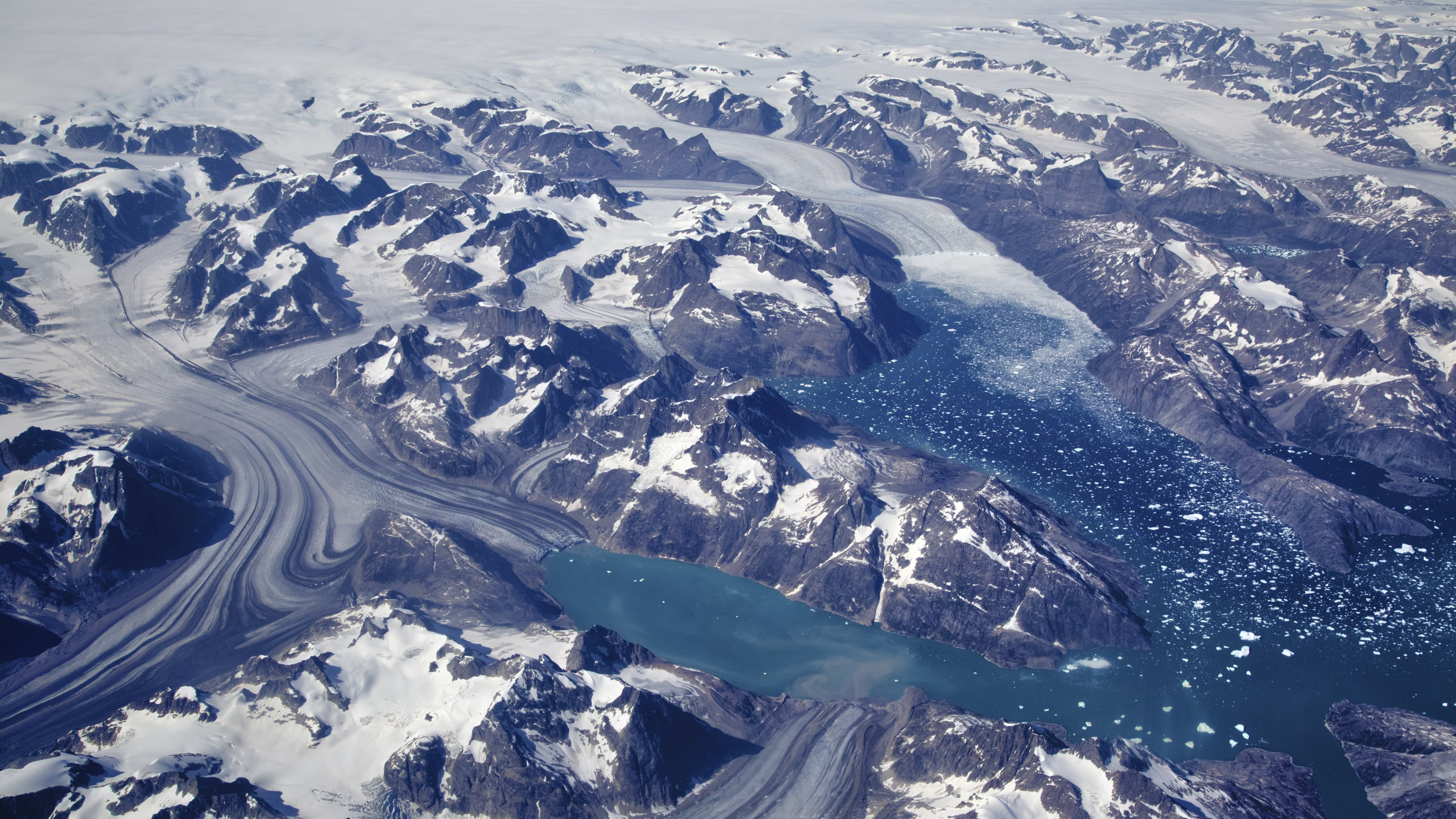 Aerial view of snow-covered mountains, glaciers flowing into icy blue waters with floating ice chunks.