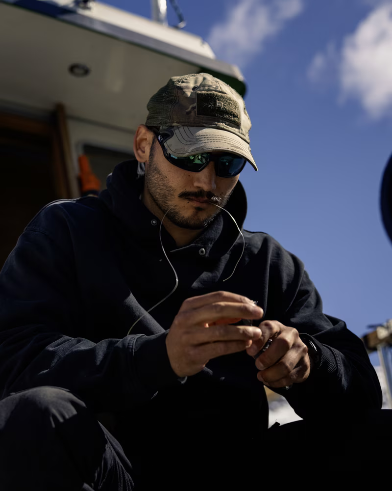 Man in sunglasses and cap focused on tying fishing line on a boat under sunny sky.
