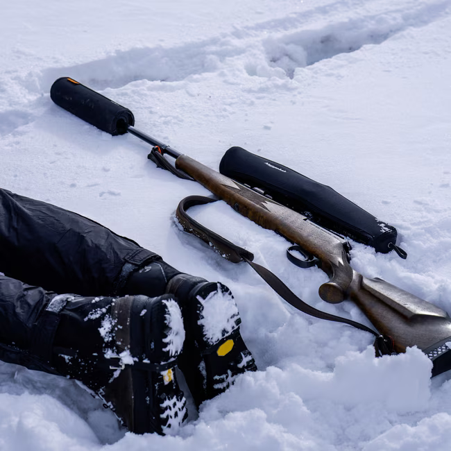 Hunting rifle with suppressor laying on snow next to person wearing black boots and pants.