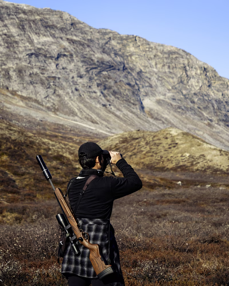 Person with a rifle on their back using binoculars in a mountainous hunting area.