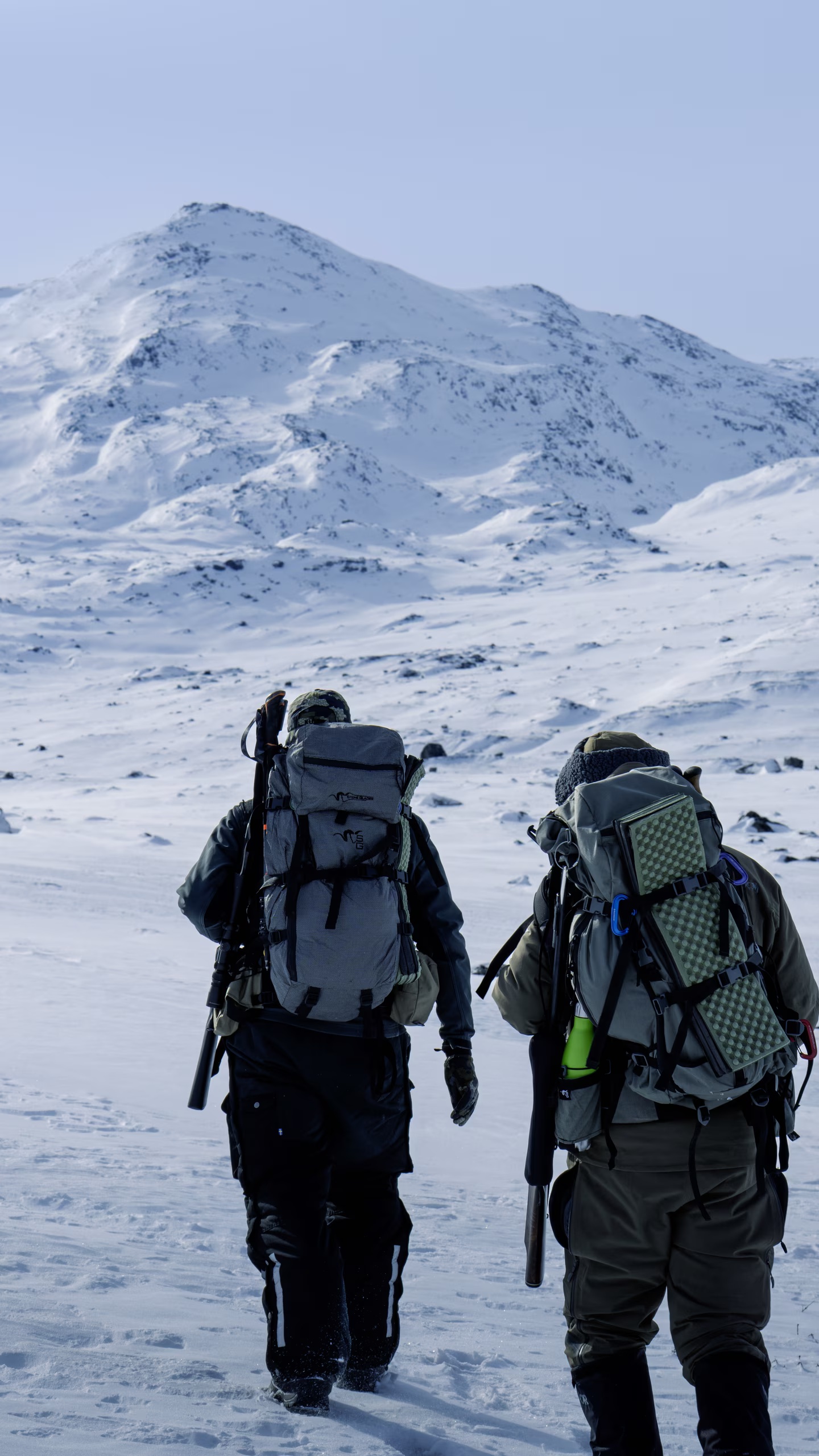 Two hikers with large backpacks and gear walking on snowy terrain toward snow-covered mountains.