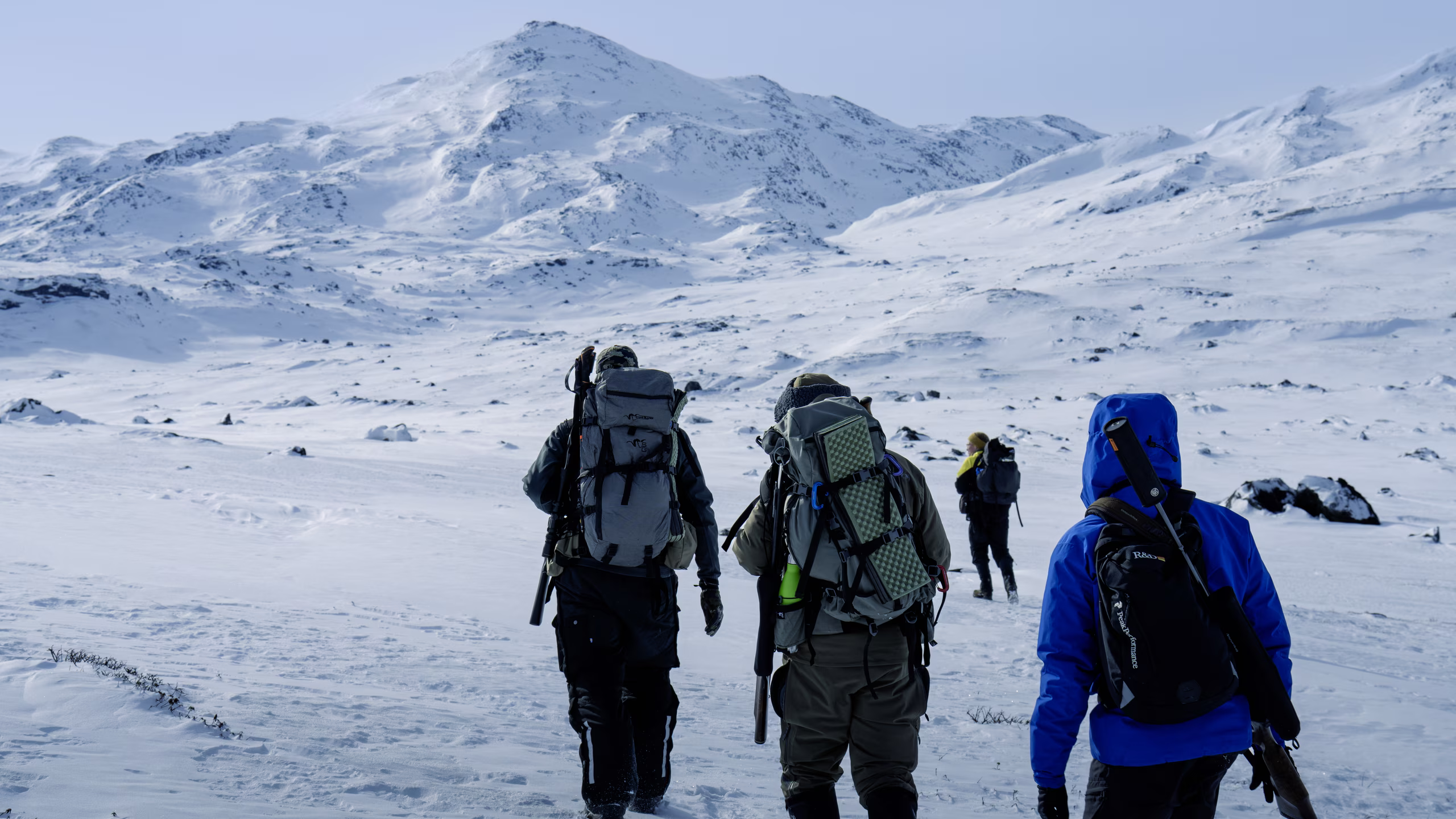 Four hunters with backpacks and rifles walk through snowy mountainous terrain under a pale sky.