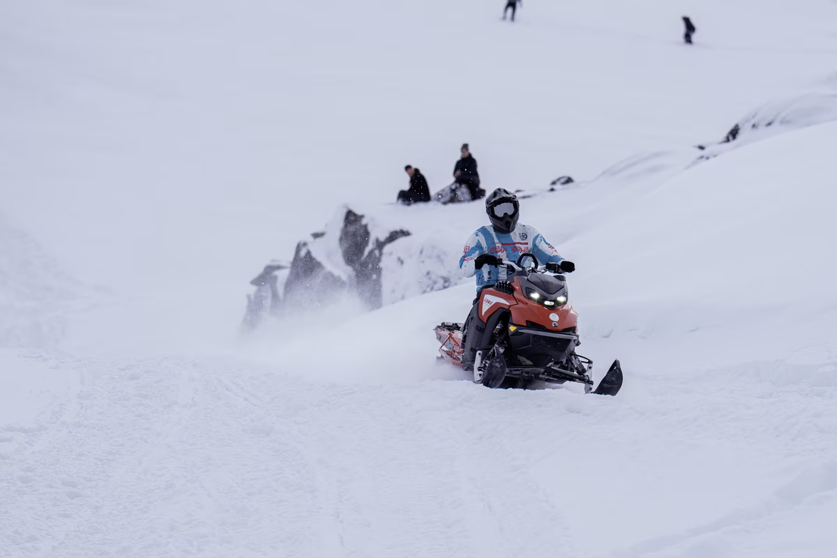 Person in helmet riding an orange snowmobile on a snowy mountain with people sitting on rocks.