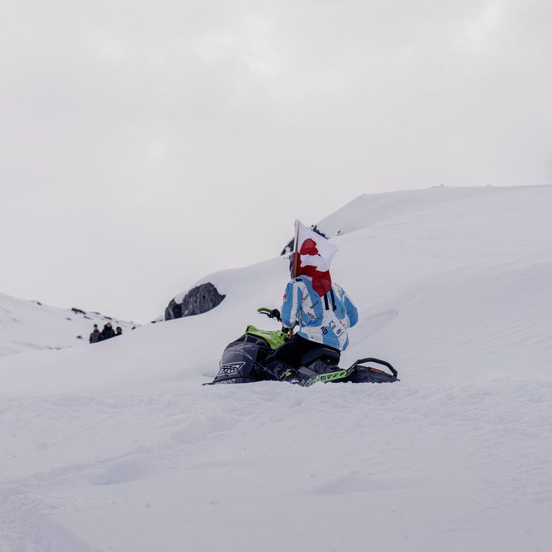 Person in winter gear with red-and-white flag on snowmobile amid snowy mountains.