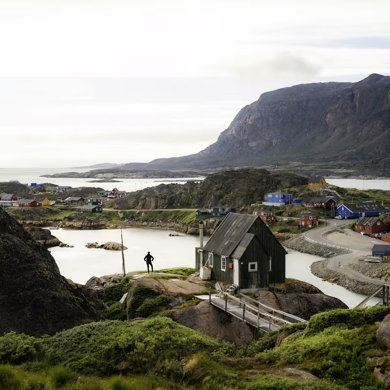 Person standing near a small cabin by water with colorful houses and mountains in the background.