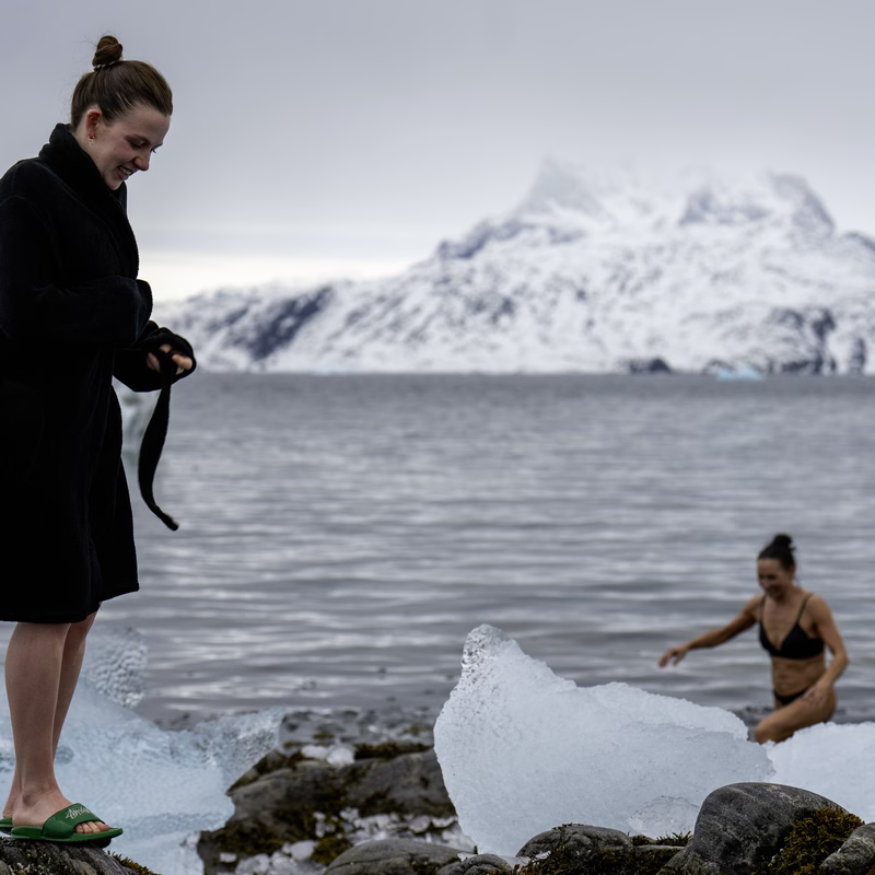 Woman in a black robe watches another woman in a swimsuit enter icy water near snow-covered mountains.