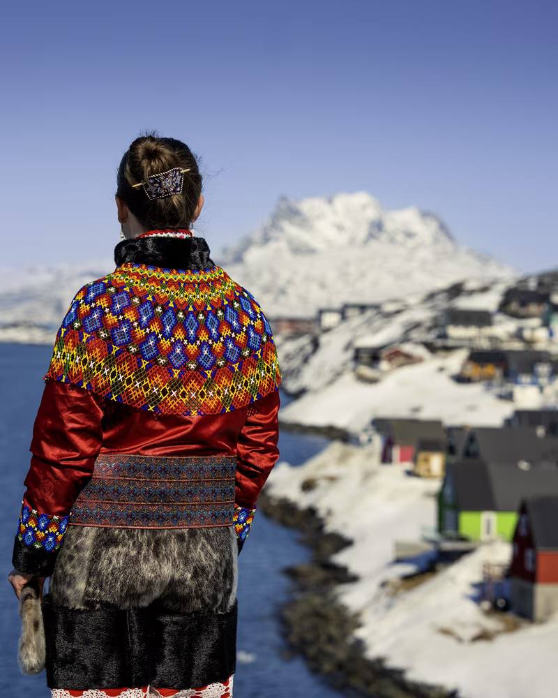 Person in colorful traditional attire standing by a snowy river with houses and mountains in the background.