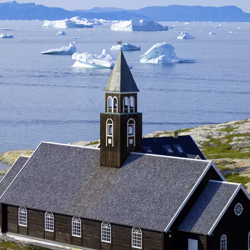 Dark wooden church on rocky shore with floating icebergs and mountains in the background.