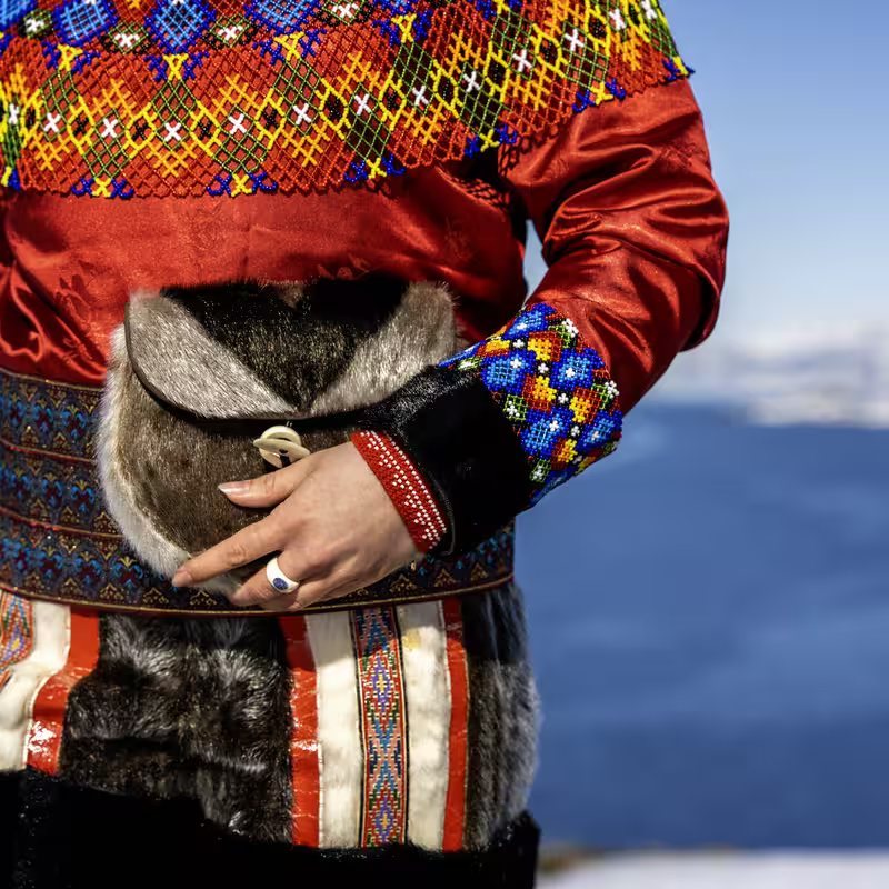 Person wearing traditional red and beaded Sami clothing with fur bag and ring on hand.