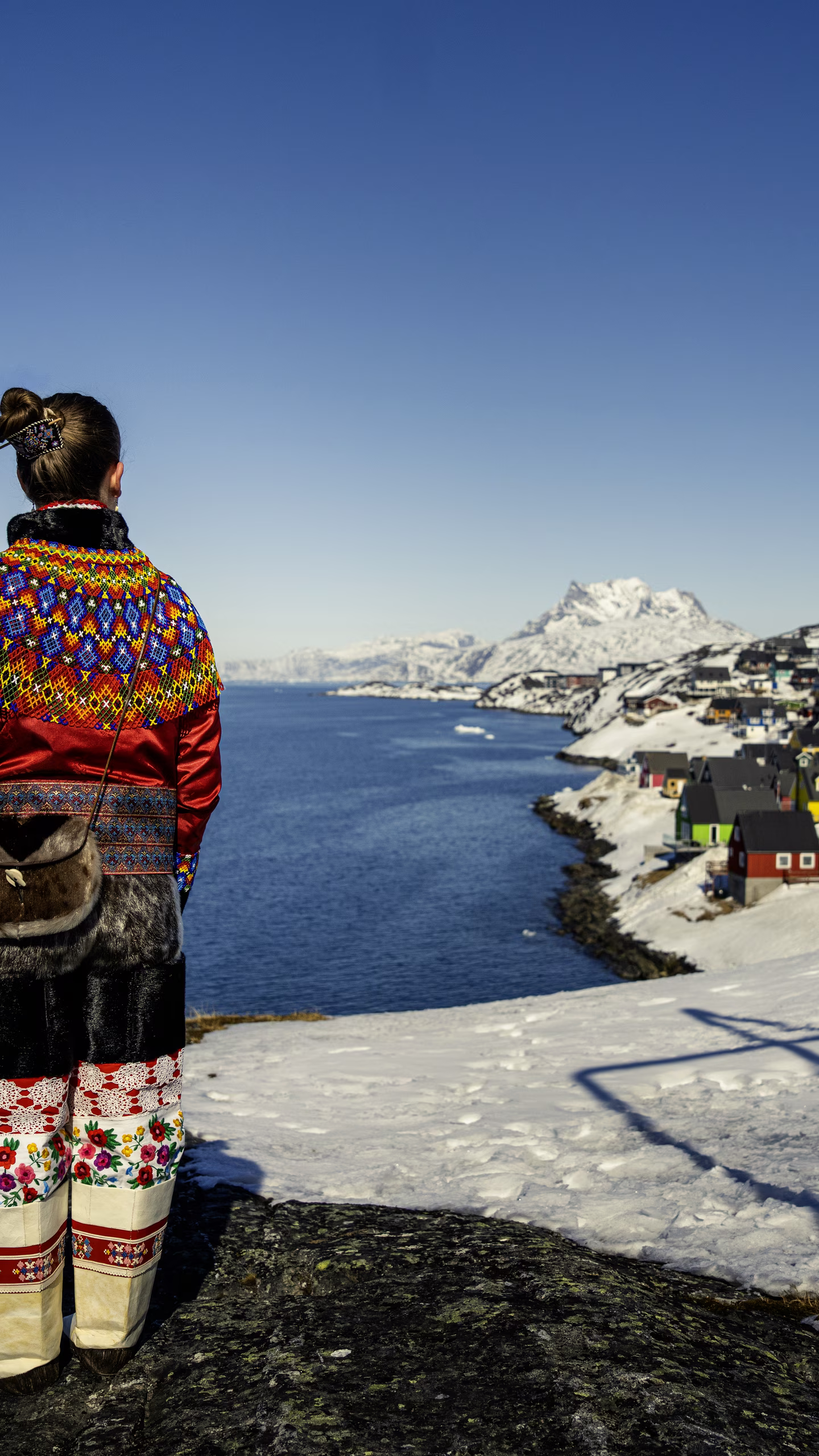 Person in colorful traditional clothing overlooking snowy village and fjord under clear blue sky.