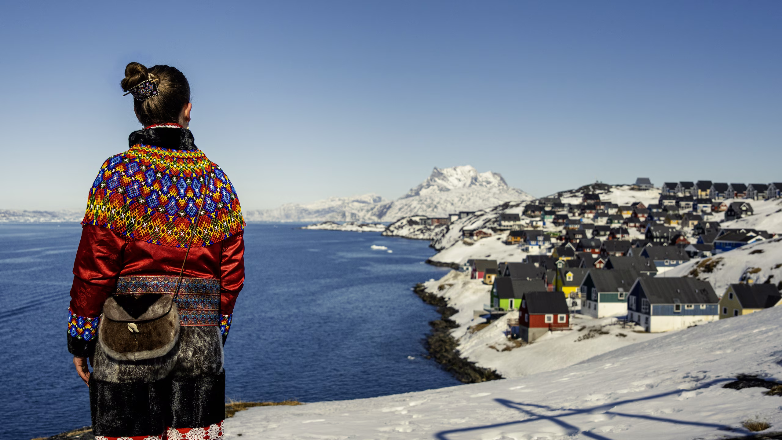 Person in colorful traditional clothing overlooks snowy coastal village and calm sea under clear sky.