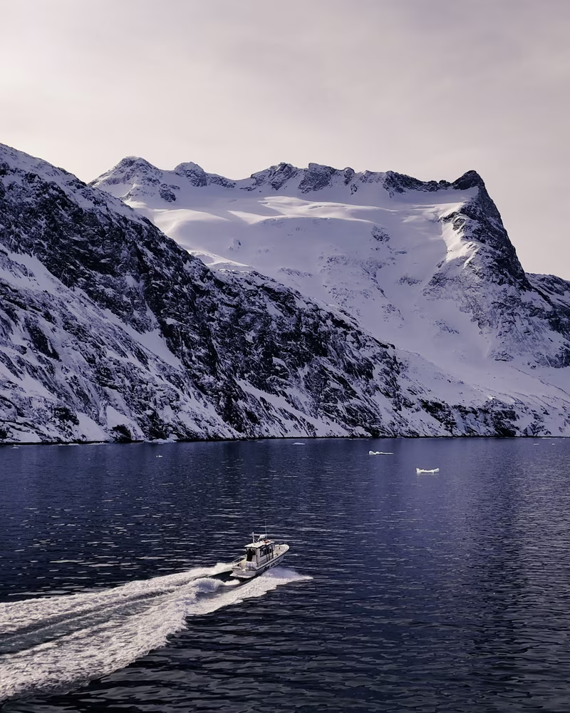 Small boat speeding on a calm blue fjord with snowy mountains in the background under a pale sky.