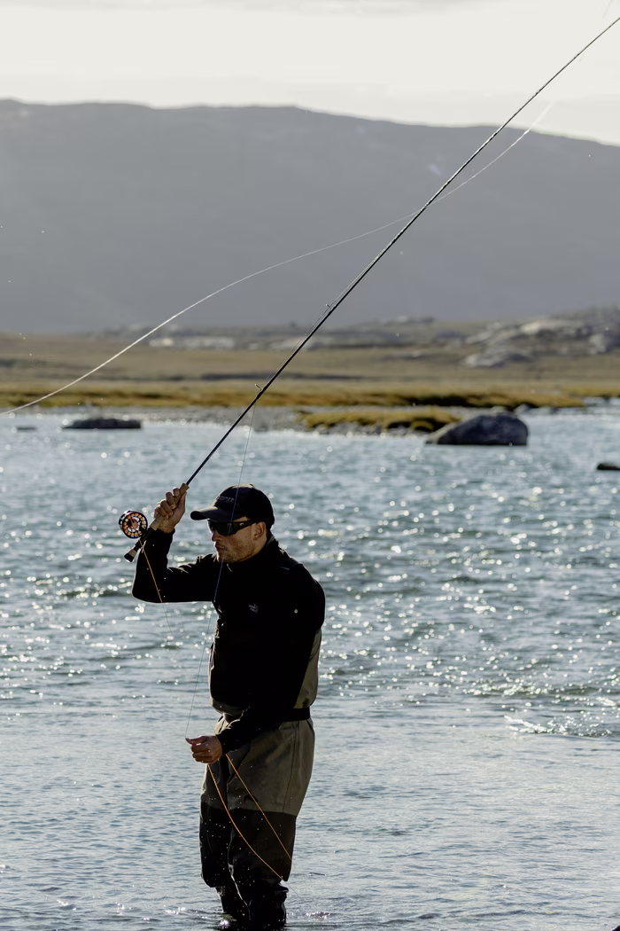 Man fly fishing in a river wearing waders with mountains in the background under daylight.