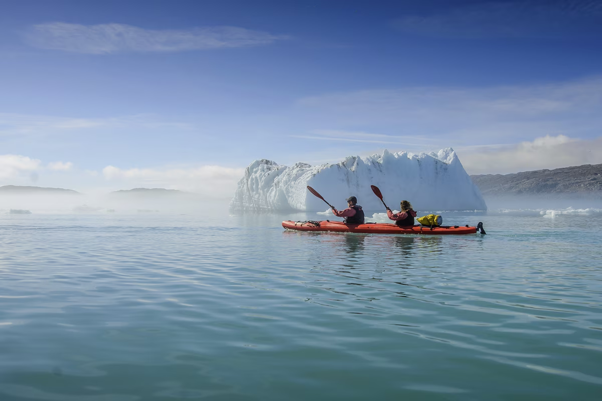 Two people kayaking on calm water near a large iceberg under a blue sky.