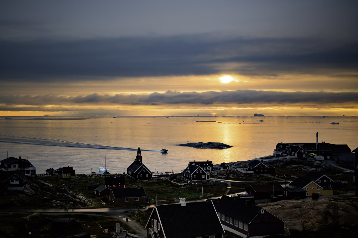 Coastal village with dark houses, a church, and boat on calm sea under sunset with dramatic clouds.