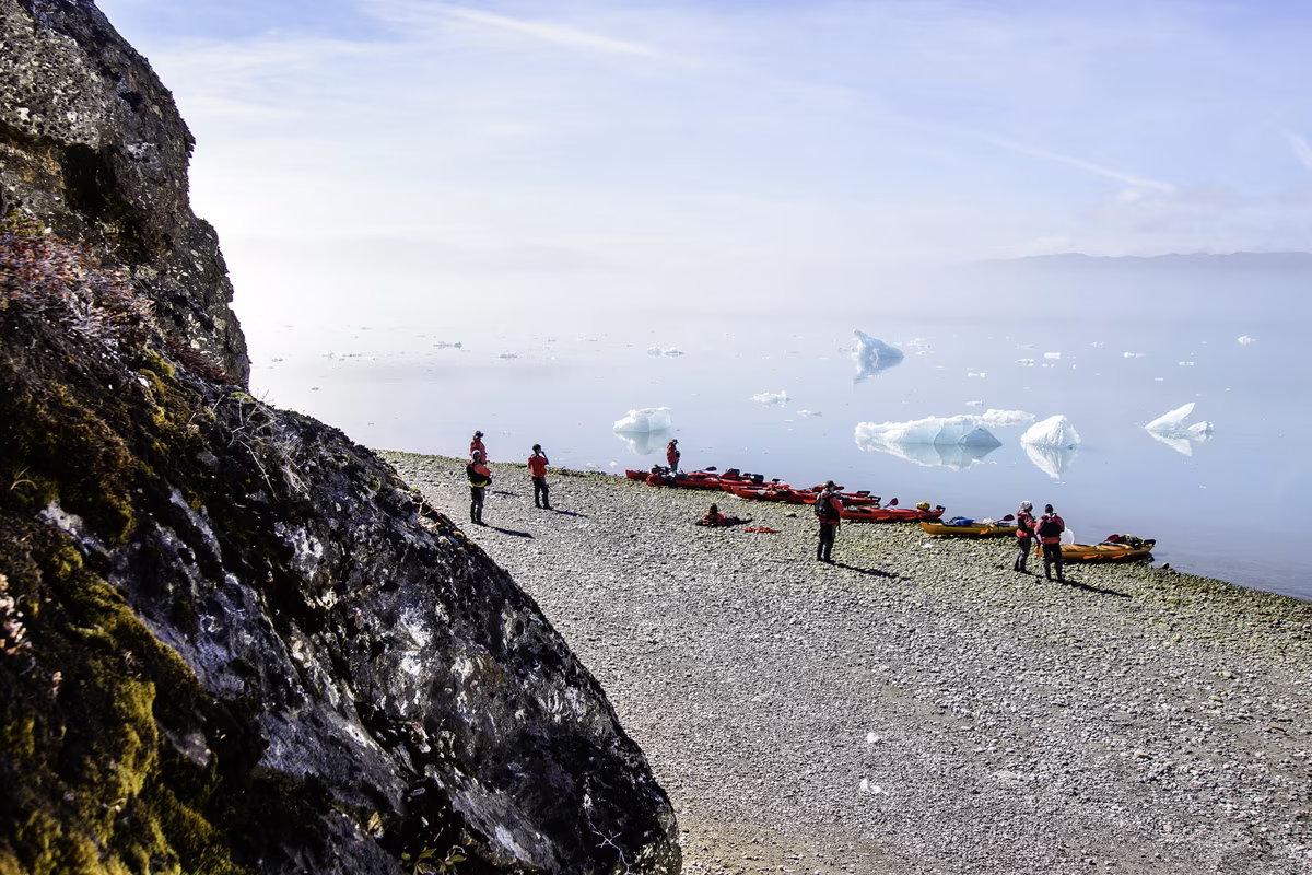 Group of people in red jackets near kayaks on a rocky shore with floating icebergs in calm water.