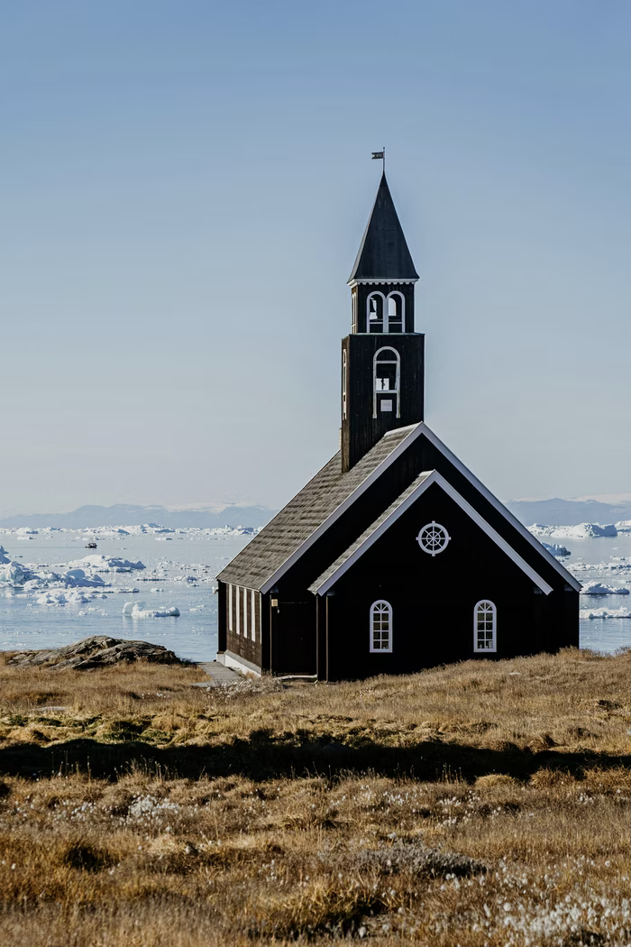 Black wooden church with white trim on grassy land near icy arctic water under clear sky.