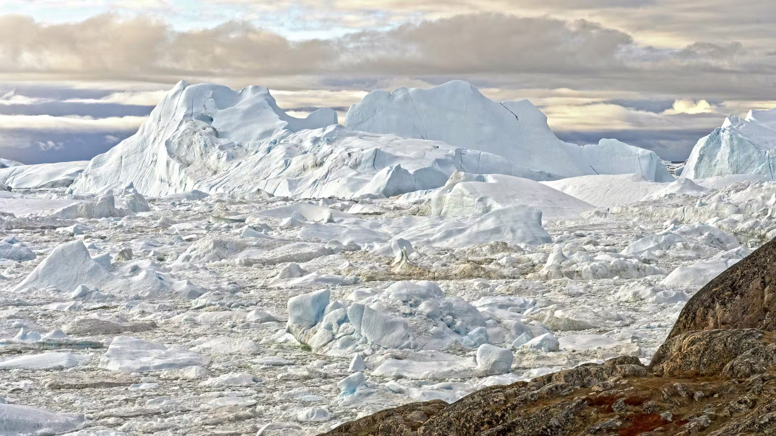 Frozen icy landscape with large icebergs and rocky foreground under a cloudy sky.