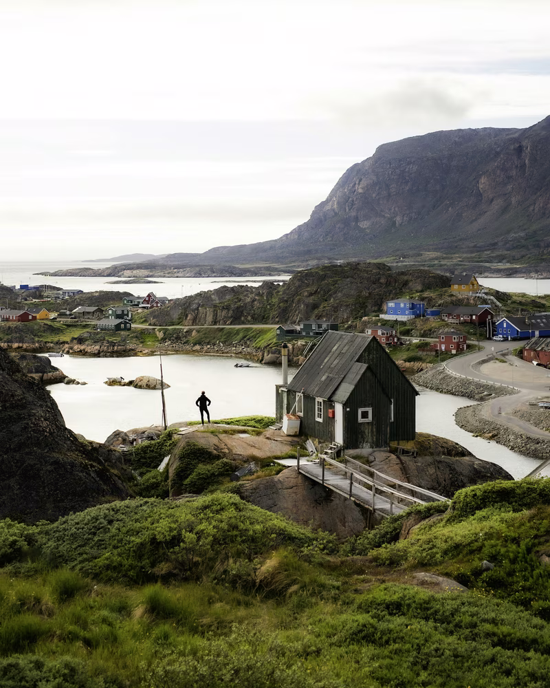 Person standing near a small house on rocky terrain with colorful houses and mountains in the background.