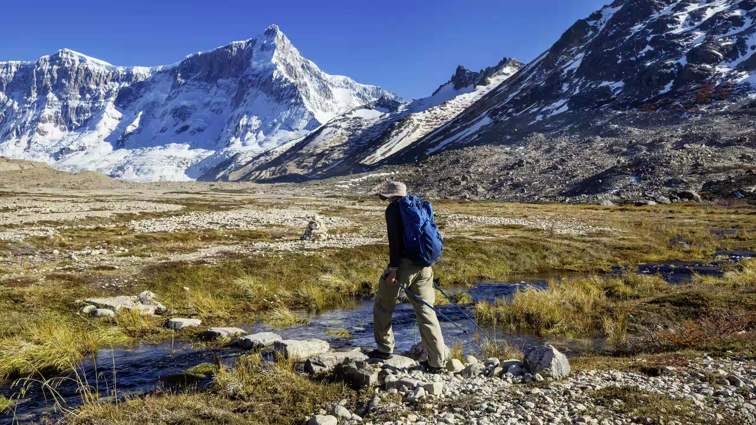 Hiker with blue backpack crossing rocky stream in a mountainous landscape with snow-capped peaks.