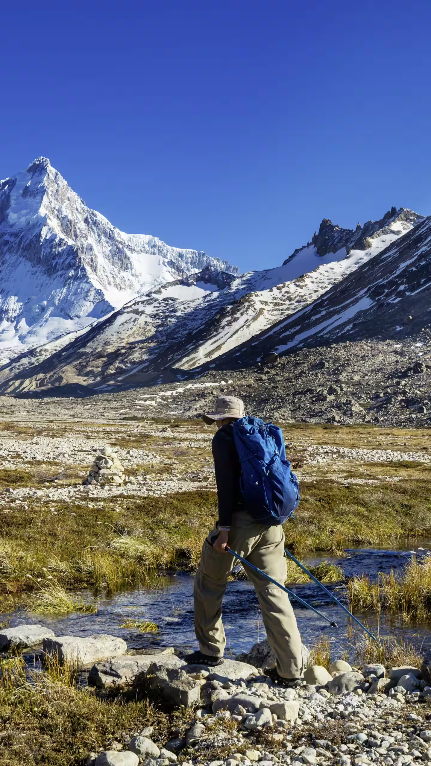 Hiker with blue backpack crossing rocky stream in a mountain valley with snowy peaks under clear sky.