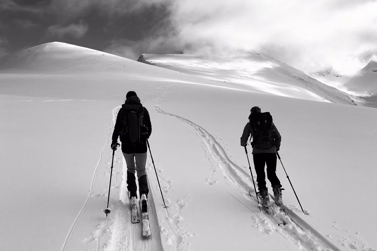 Two skiers with backpacks skiing uphill on a snowy mountain under cloudy sky.