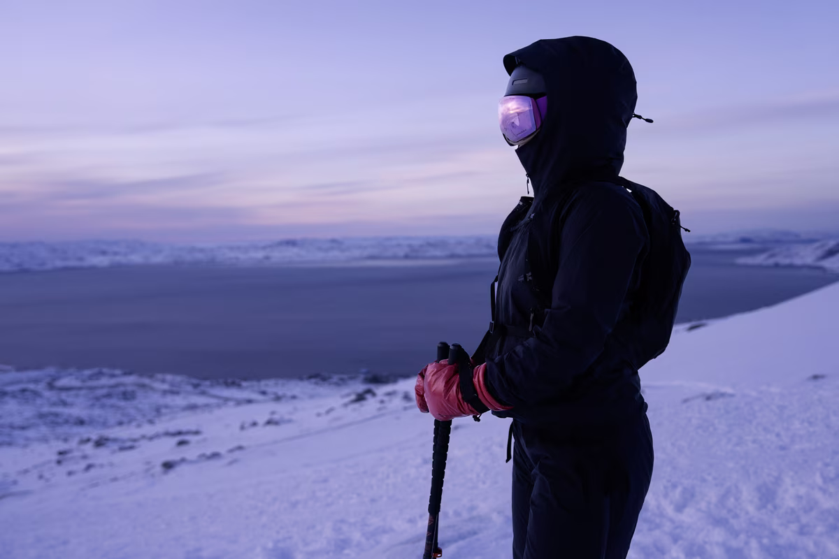 Person in black winter gear and pink gloves holding poles overlooking snowy landscape at dusk.
