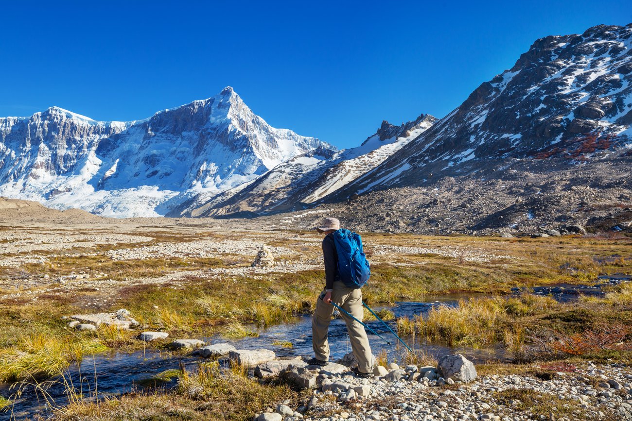 Hiker with blue backpack crossing rocky stream in a valley with snowy mountains under a clear blue sky.