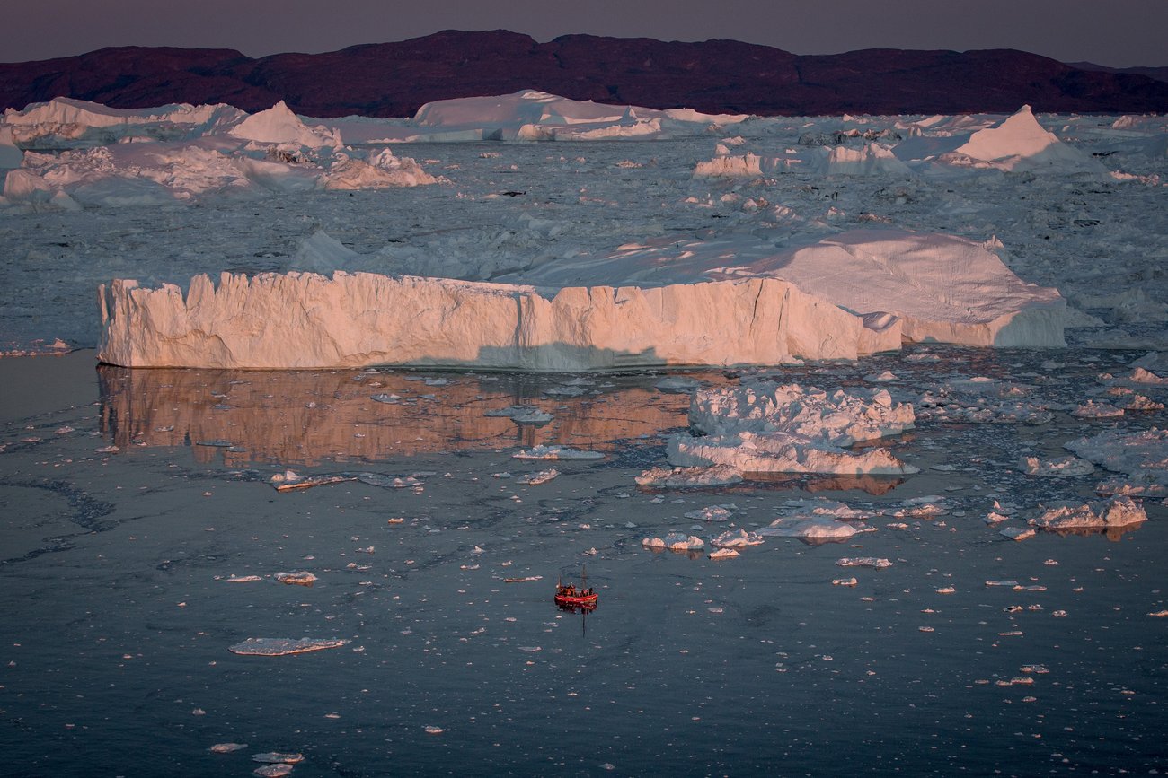 Small red boat surrounded by chunks of ice floating near large icebergs at dusk with mountains in background.