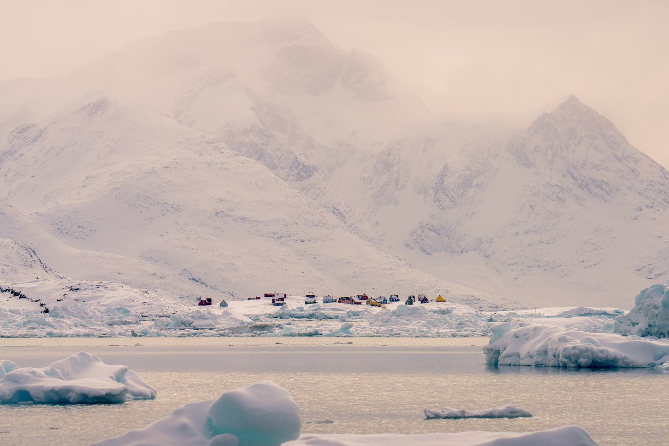 Small colorful houses on snowy shore with icy water and large snow-covered mountains in the background.