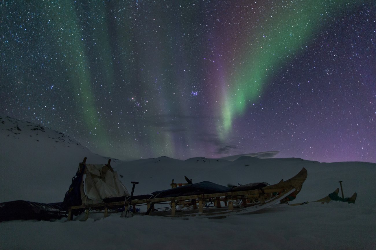 Northern lights glowing above snowy mountains and a wooden sled resting on the snow.