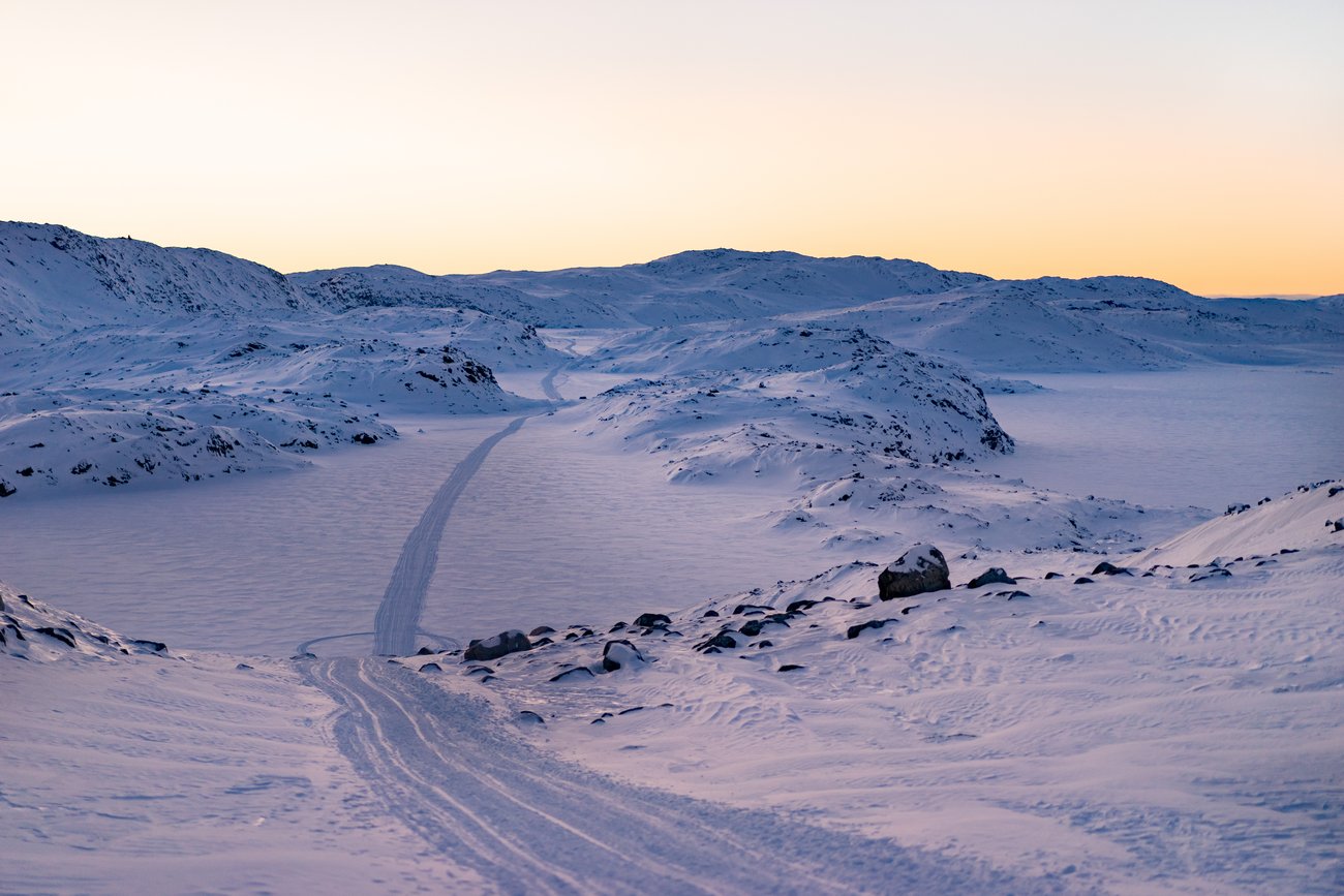 Snow-covered mountainous landscape with a narrow snow trail leading through the valley at sunset.