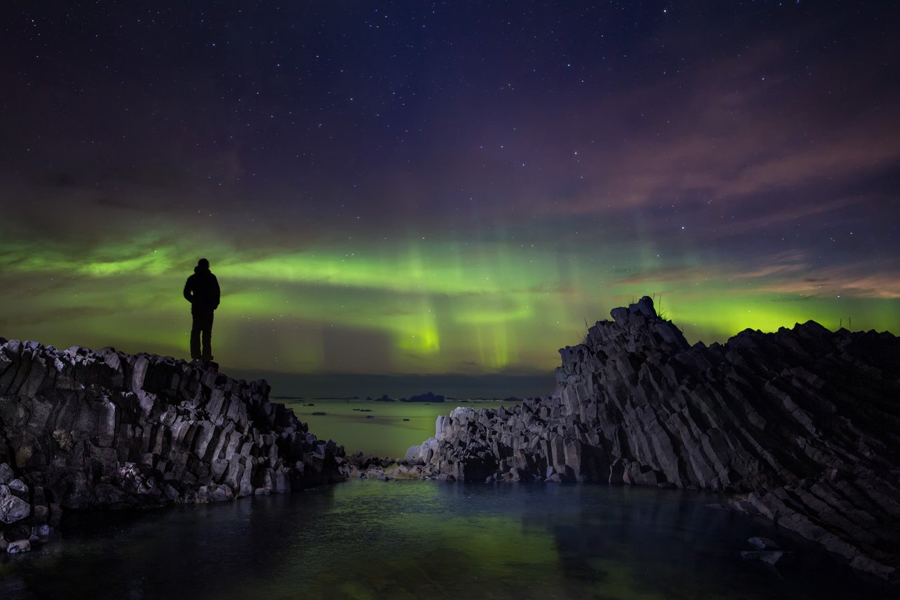 Silhouette of person on rocky shore watching green northern lights under starry night sky.