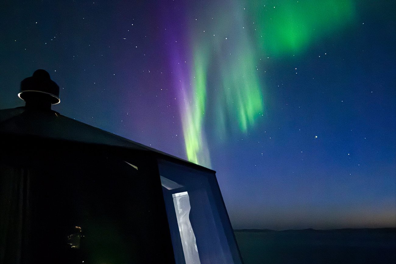 Aurora borealis in green and purple hues lighting up a starry night sky above a dark cabin window.