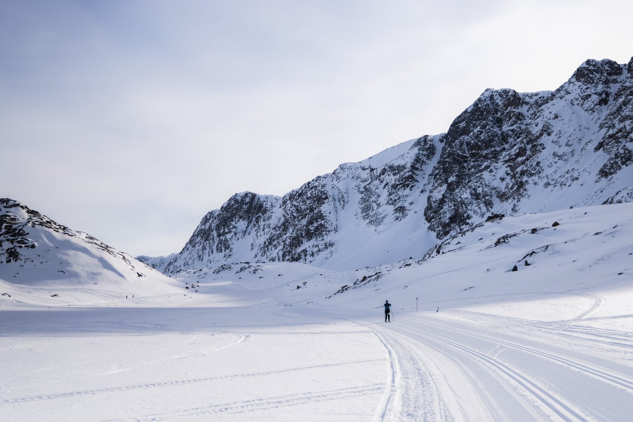 Skier on groomed trail in snowy mountainous landscape under pale sky.