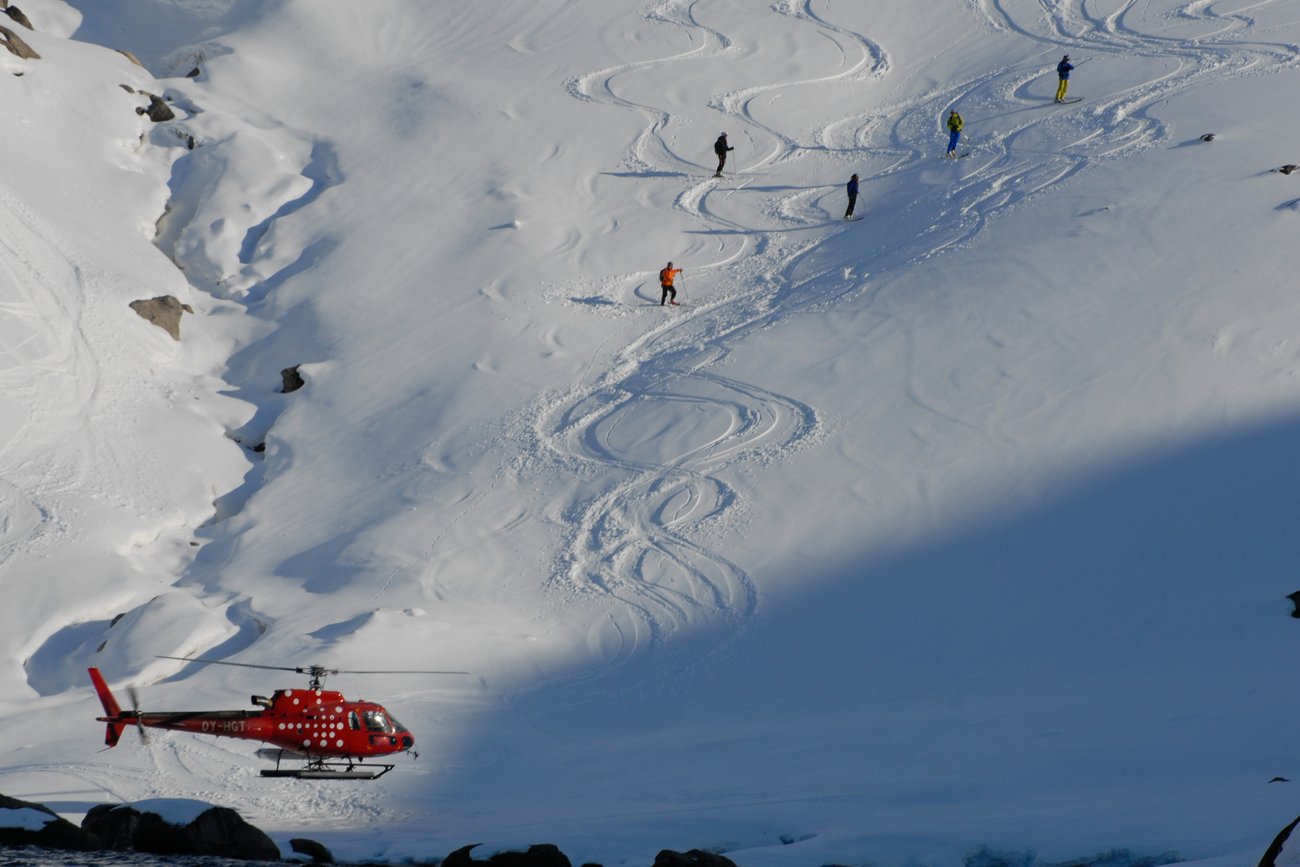 Red helicopter near snowy mountainside with four skiers standing on ski tracks in fresh snow.