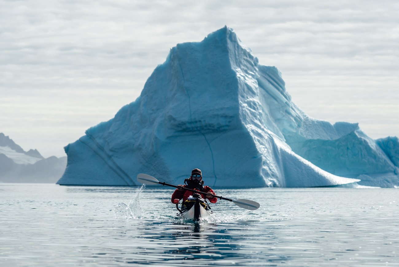 Person kayaking on calm water with a large blue iceberg in the background under cloudy sky.