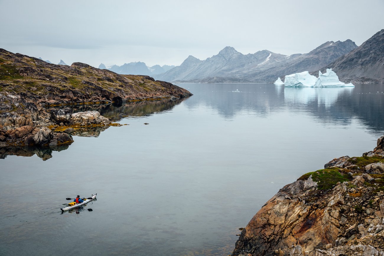 Person kayaking in calm water near rocky shores with icebergs and mountains in the distance.