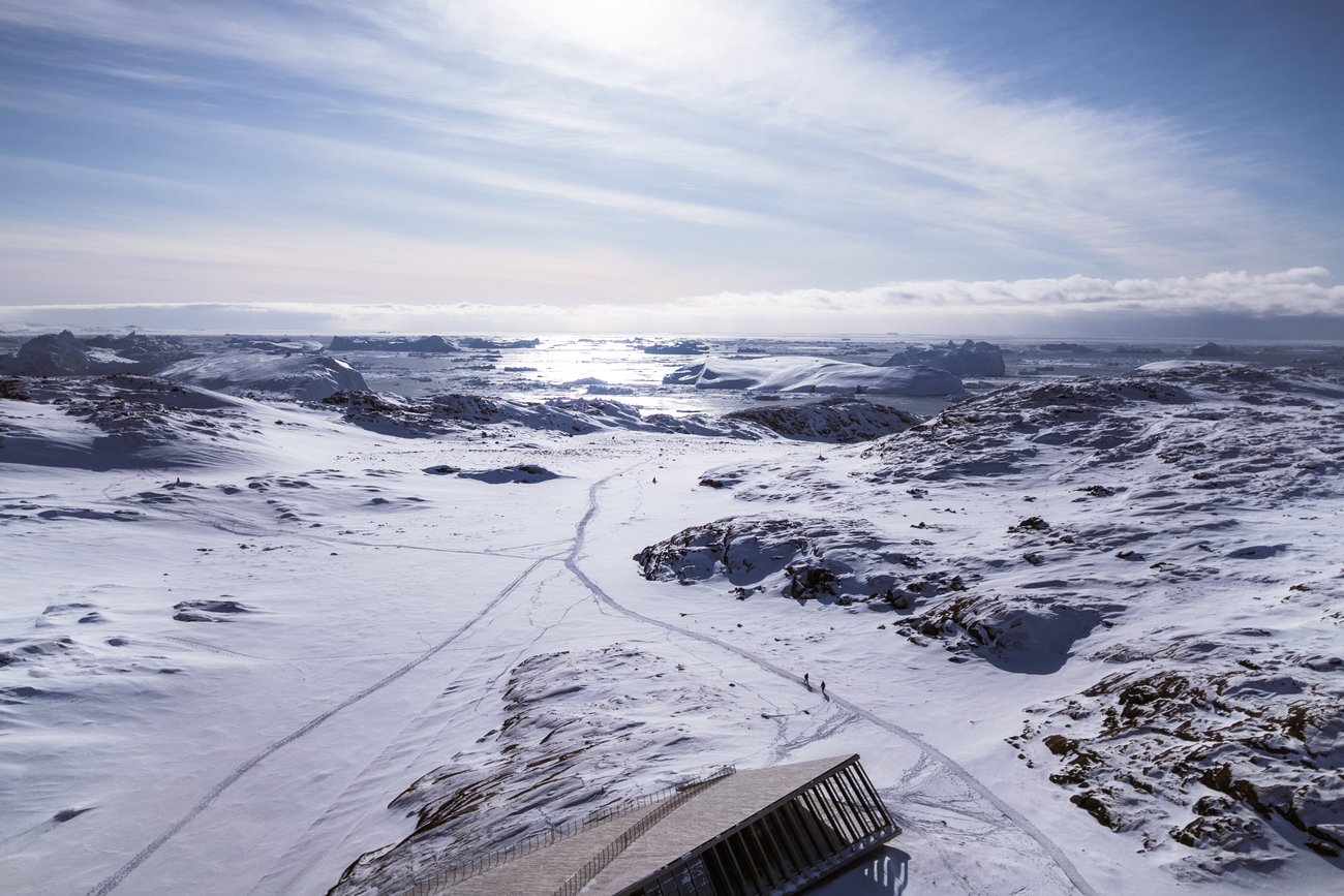 Snowy Arctic landscape with rocky hills, distant icebergs, and two small people walking near a wooden structure.