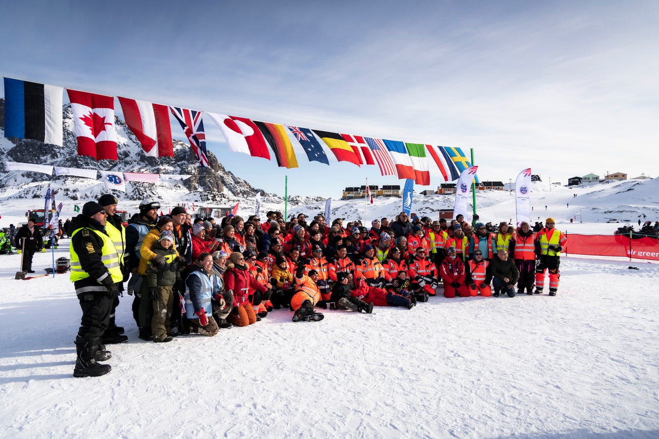 Large group of people in winter gear posing in snow with flags and snowy mountains in the background.