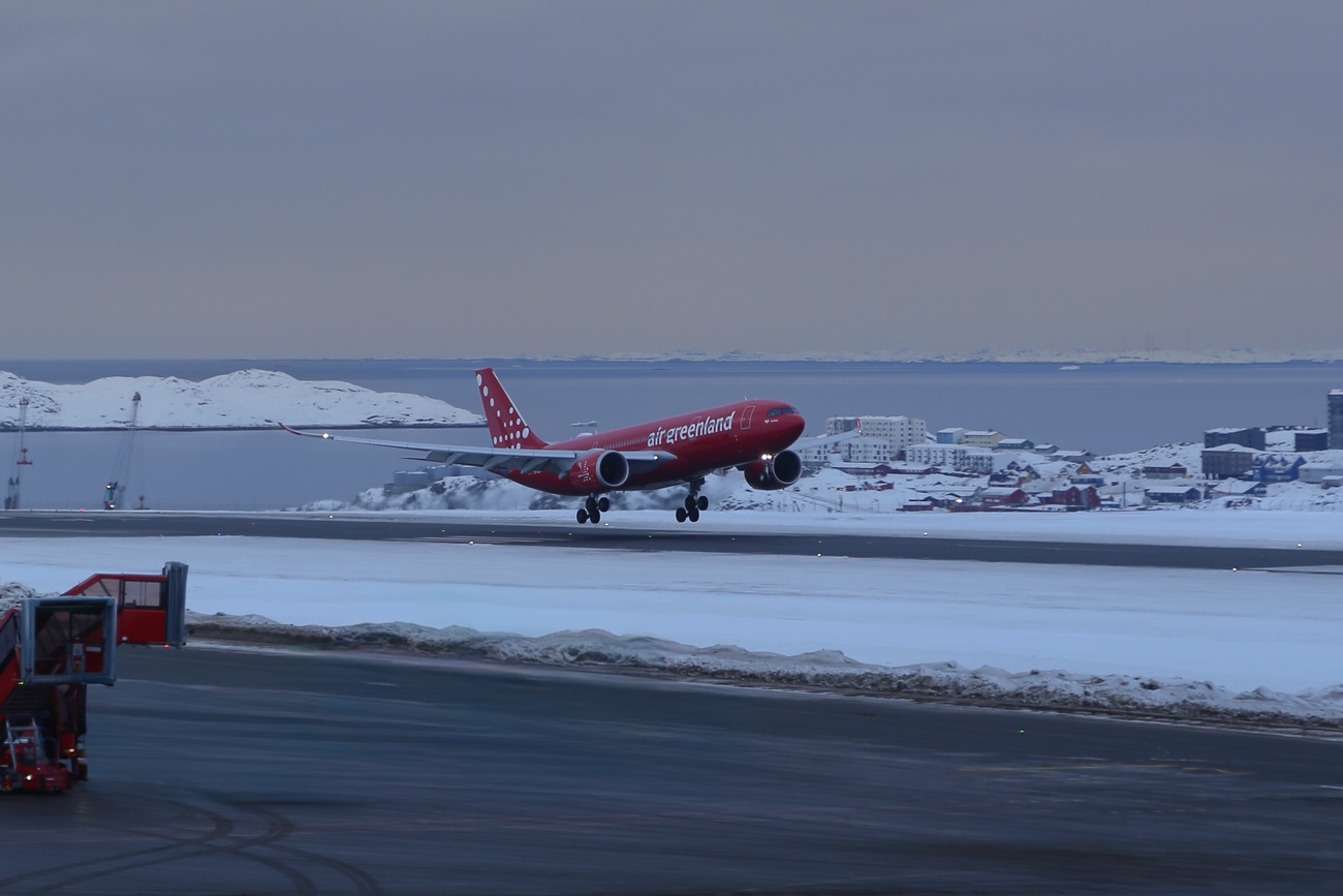 Red Air Greenland plane taking off from snowy runway with a snowy town and sea in the background.