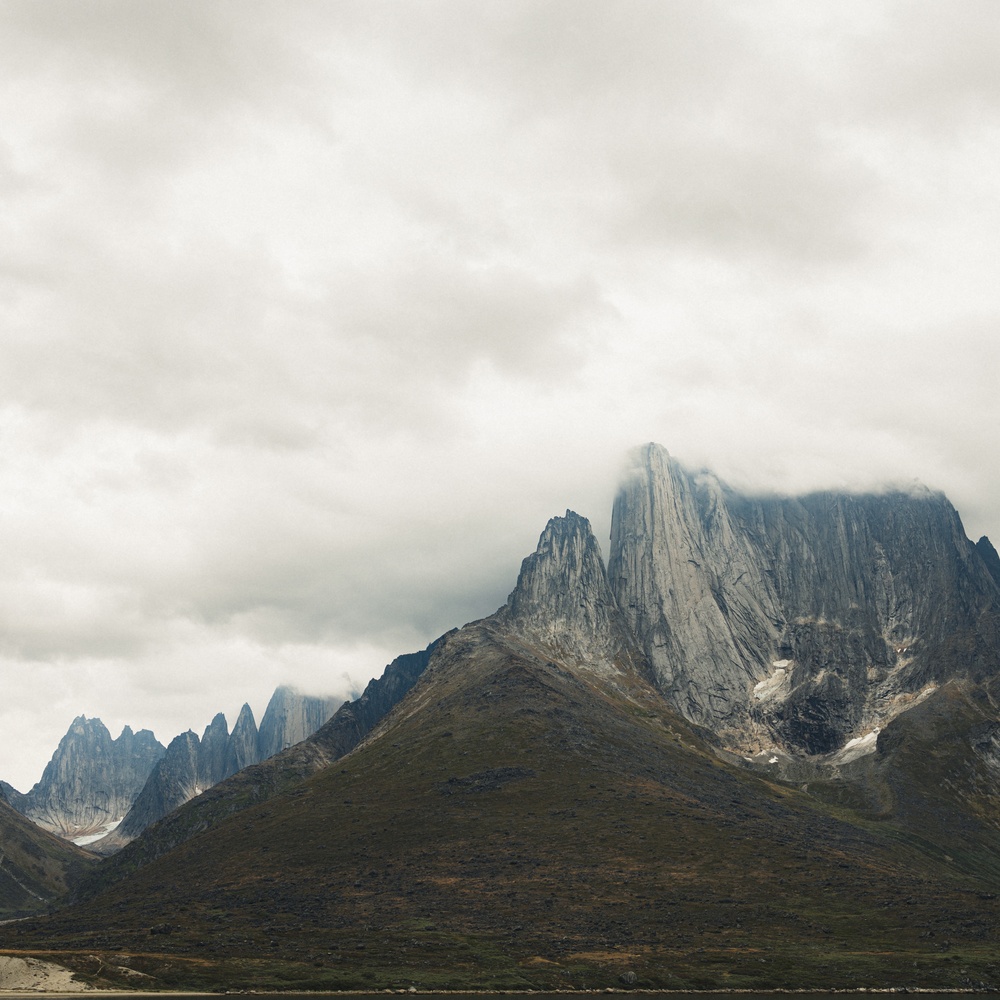 Jagged Tasermiut mountain peaks partly shrouded in clouds under a gray overcast sky.