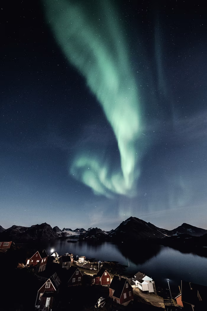 Northern lights glowing green above a small village with mountains and water at night.