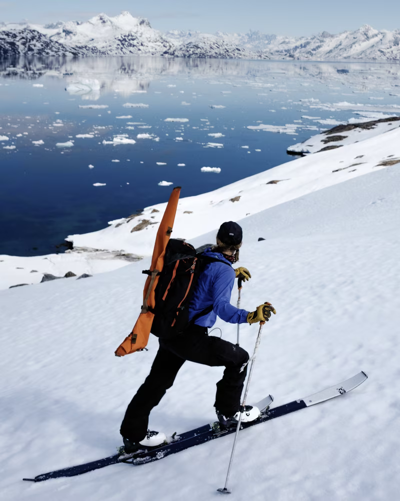 Skier ascending snowy slope with ski poles near icy water and snow-covered mountains.