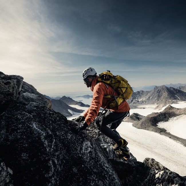 Climber wearing helmet and orange jacket scaling rocky mountain with snowy peaks in the background.
