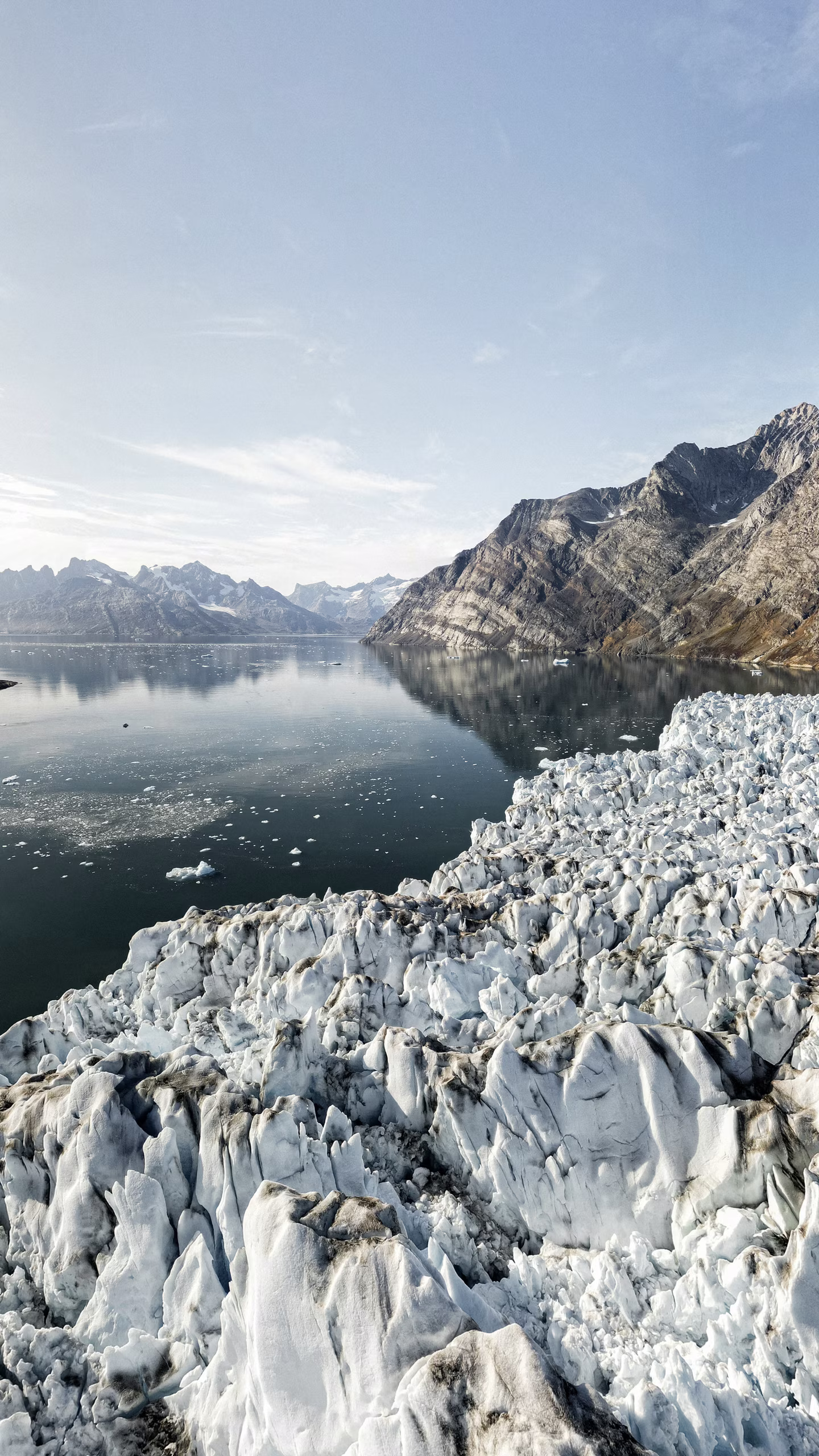 Jagged glacier edges meeting calm water beside rocky mountains under a clear sky.